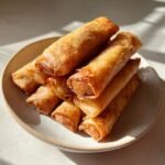 A stack of freshly fried, golden brown Lumpia rolls piled high on a light ceramic plate.