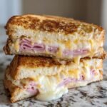 Close-up of a perfectly grilled Ham And Cheese Sandwich cut in half and stacked, showing melted cheese oozing out.