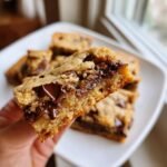 A hand holding up a thick, gooey bar cut from Sheet Pan Chocolate Chip Cookies, showing melted chocolate chips.