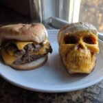 A double cheeseburger sits next to a bun top decorated with melted cheese to look like a skull, representing Halloween Cheeseburger Skulls.