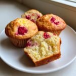 Close-up of a plate showing three whole Lemon Raspberry Muffins and one cut in half, revealing the moist interior.