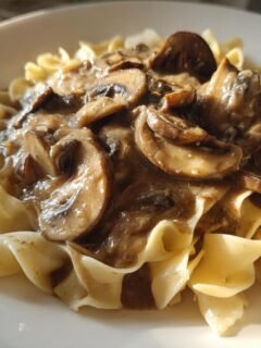 Close-up of creamy Mushroom Stroganoff generously spooned over wide egg noodles on a white plate.