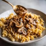 A spoonful of creamy Mushroom Stroganoff sauce with sliced mushrooms is being poured over a bed of egg noodles.