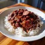A hearty bowl of Red Beans And Rice topped with slices of smoked sausage, served on a white plate.