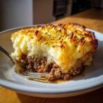 A close-up of a serving of Shepherds Pie with a fork digging into the rich meat filling beneath the golden, crispy mashed potato topping.