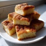 A stack of six square Snickerdoodle Bars topped generously with cinnamon sugar, resting on a white plate.