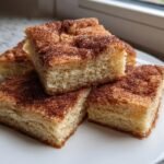 Four square cuts of soft Snickerdoodle Bars, topped generously with cinnamon sugar, stacked on a white plate.