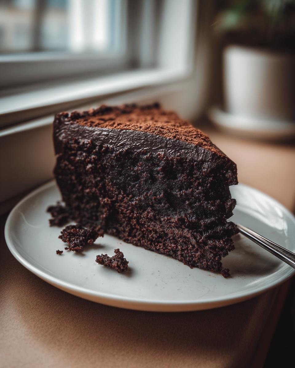 A dark, moist slice of Chocolate Doom Cake dusted with cocoa powder on a white plate.