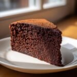 A moist, dark slice of Chocolate Doom Cake dusted lightly with cocoa powder, sitting on a white plate.