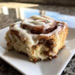 Close-up of a single, gooey Christmas Cinnamon Rolls slice topped with thick white icing on a white plate.