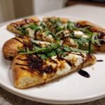 Close-up of a slice of Fig Jam Goat Cheese Pizza topped with fresh arugula and balsamic glaze.