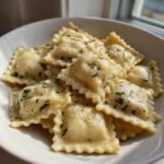 A close-up of a white bowl filled with cheesy Italian Ravioli topped with melted butter sauce and fresh chopped parsley.