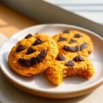 Three orange pumpkin-shaped cookies decorated like Jack O Lanterns with dark chocolate faces. One cookie has a bite taken out.