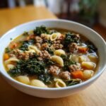 Close-up of a white bowl filled with hot Sausage Kale Soup, featuring crumbled sausage, dark green kale, pasta shells, and carrots in broth.