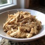 A close-up of perfectly shredded Crockpot Chicken piled high in a white bowl, glistening with juices.