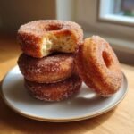 A stack of three golden brown Apple Cider Donuts coated in cinnamon sugar, with the top one having a bite taken out.