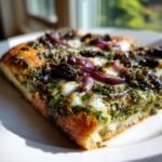 Close-up of a thick slice of Witch Pesto Pizza topped with melted mozzarella, red onion, and dark toppings.