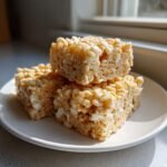Three golden brown squares of homemade Rice Krispie Treats stacked on a white plate.