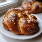 Close-up of a freshly baked, glossy Pretzel Bread, braided and topped with coarse pearl sugar, sitting on a white plate.
