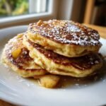 A close-up of three fluffy Apple Pie Pancakes stacked on a white plate, topped with powdered sugar and cinnamon apples.