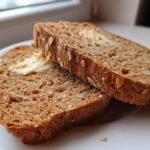 Two slices of toasted Ezekiel Bread, lightly spread with melting butter, resting on a white plate near a window.
