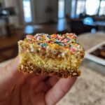 A hand holding a thick, moist slice of Cake Batter Bars topped with white glaze and colorful sprinkles.