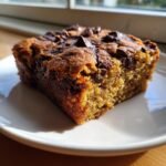 A close-up, sunlit shot of a single slice of moist Chocolate Chip Banana Bars on a white plate.