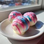 Three white chocolate-coated Cotton Candy Cheesecake Bombs drizzled with bright pink and blue icing and sprinkles, sitting on a white plate.