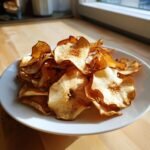 A pile of crispy, baked Apple Chips dusted with cinnamon served on a white plate near a sunny window.