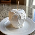 A single, freshly made Marshmallow Apples treat covered in thick, white, fluffy topping, resting on a white plate.