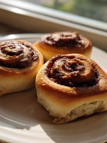 Three golden-brown Nutella Rolls with gooey centers sitting on a white plate near a sunny window.