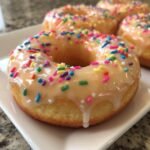 A close-up of a freshly glazed Funfetti Donut topped with colorful sprinkles, sitting on a white plate.