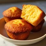 Close-up of golden Cornbread Cupcakes stacked in a bowl, one broken open showing the moist interior.
