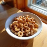 A white bowl filled with warm, glistening Hot Buttered Cheerios sitting on a wooden table near a sunlit window.