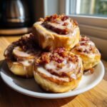 A stack of four delicious Maple Bacon Cinnamon Rolls, drizzled with white icing and topped with crispy bacon bits.