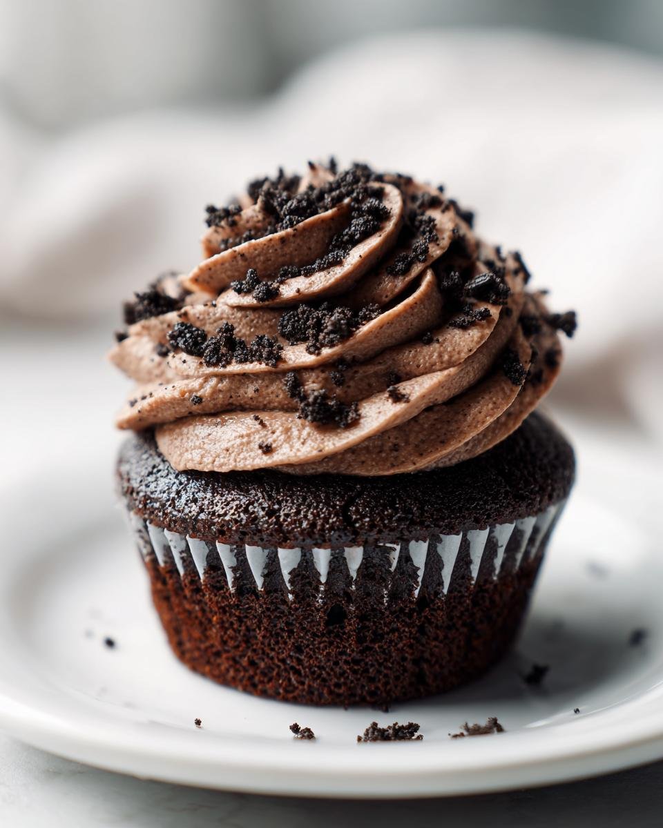 A single, rich chocolate Oreo Cupcakes topped with swirled chocolate frosting and crushed cookie pieces, sitting on a white plate.