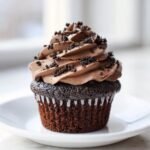 A close-up of a rich chocolate Oreo Cupcakes topped with light brown, swirled frosting and crushed cookie pieces.