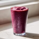 A tall glass filled with a thick, rich purple Greek Yogurt Smoothies, sitting on a white marble surface near a window.