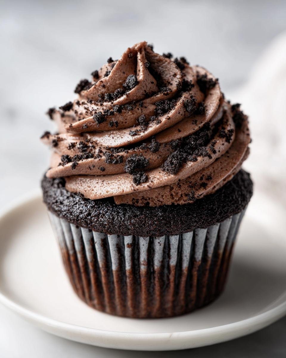 A close-up of a decadent Oreo Cupcakes featuring dark chocolate cake and swirled chocolate frosting topped with cookie crumbs.