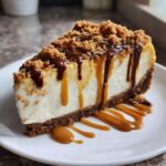 A perfect slice of Biscoff Cheesecake topped with cookie crumbs, caramel, and chocolate drizzle on a white plate.