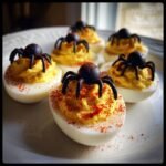 A plate of festive Spider Deviled Eggs, topped with black candy spiders and sprinkled with paprika.