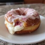 A close-up of a glazed Strawberry Cheesecake Donut, split open to show creamy white filling and pink strawberry bits.