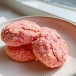 A close-up of three bright pink Strawberry Sugar Cookies stacked slightly on a light-colored plate near a window.