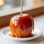 A close-up of a glossy, dripping Vegan Caramel Apples resting on a small white plate.