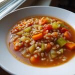 A close-up of a white bowl filled with steaming Vegetable Barley Soup featuring plump barley grains, diced carrots, and celery in a rich broth.