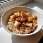 Close-up of a white bowl filled with creamy Apple Cinnamon Oatmeal, topped with diced apples and a generous dusting of cinnamon.
