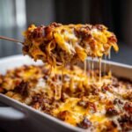 A spoonful of cheesy Bacon Cheeseburger Pasta Bake being lifted from the baking dish, showing long cheese pulls.