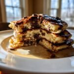 A stack of three fluffy Chocolate Chip Pancakes, cut open to show melted chocolate chips inside, drizzled with syrup.