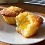 Two golden Lemon Curd Muffins on a white plate, one cut open revealing the bright yellow lemon curd center.