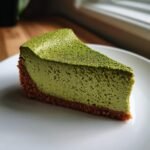 A perfect slice of vibrant green Matcha Cheesecake with a dark crust, dusted heavily with matcha powder.
